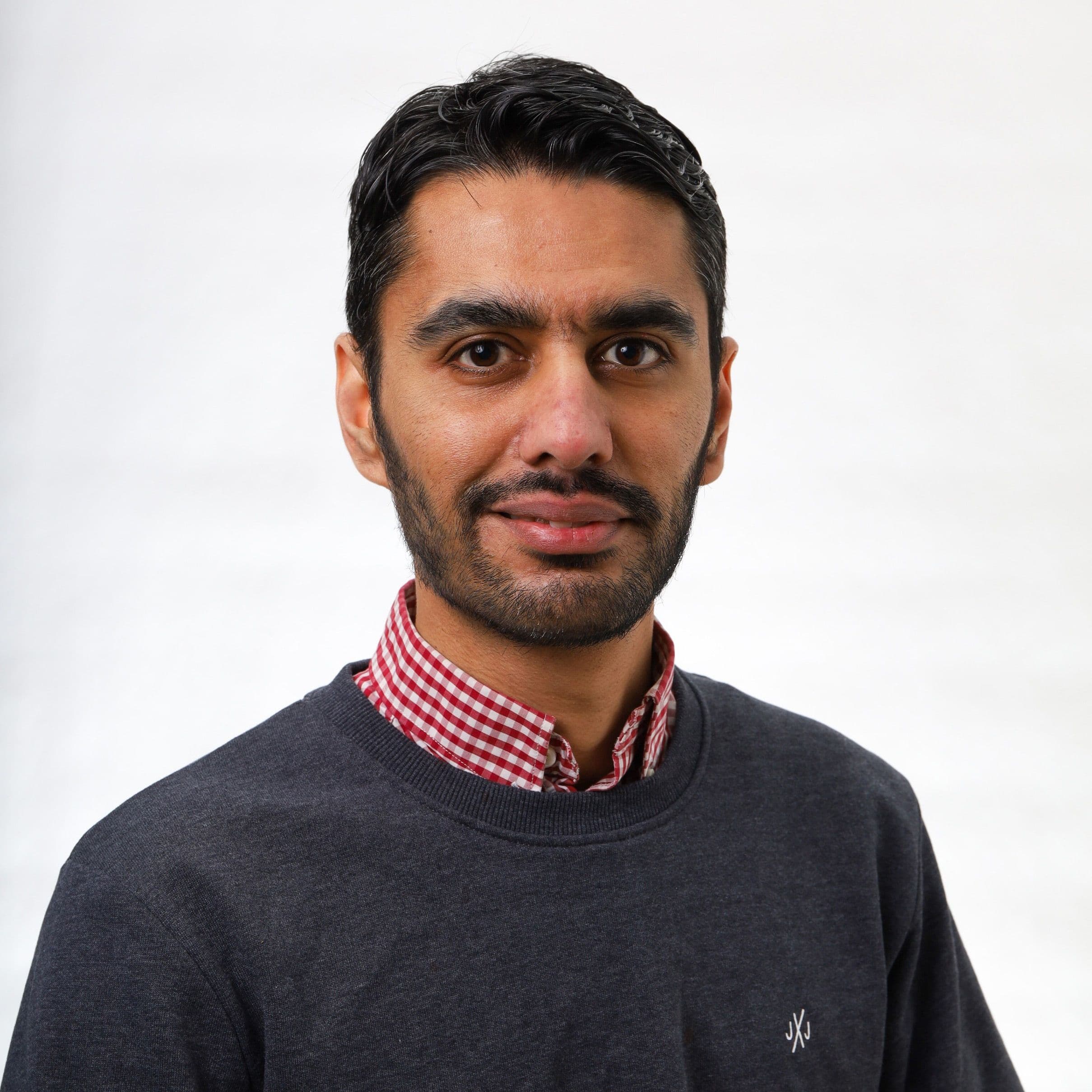 Danial smiler med mørkt hår og skjegg i rutete skjorte og mørk genser. Foto.